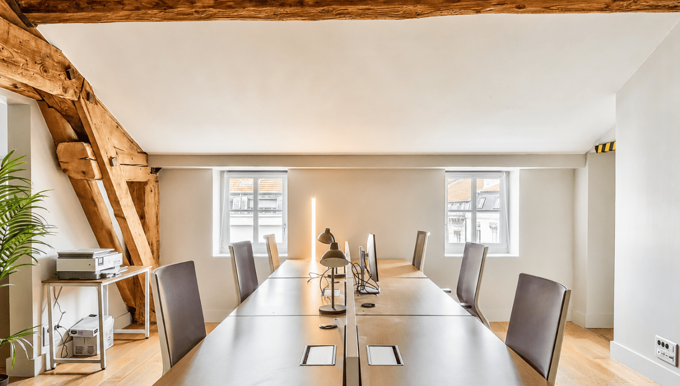 Bureau partagé avec longue table équipée d’écrans, plusieurs sièges et poutres en bois apparentes.