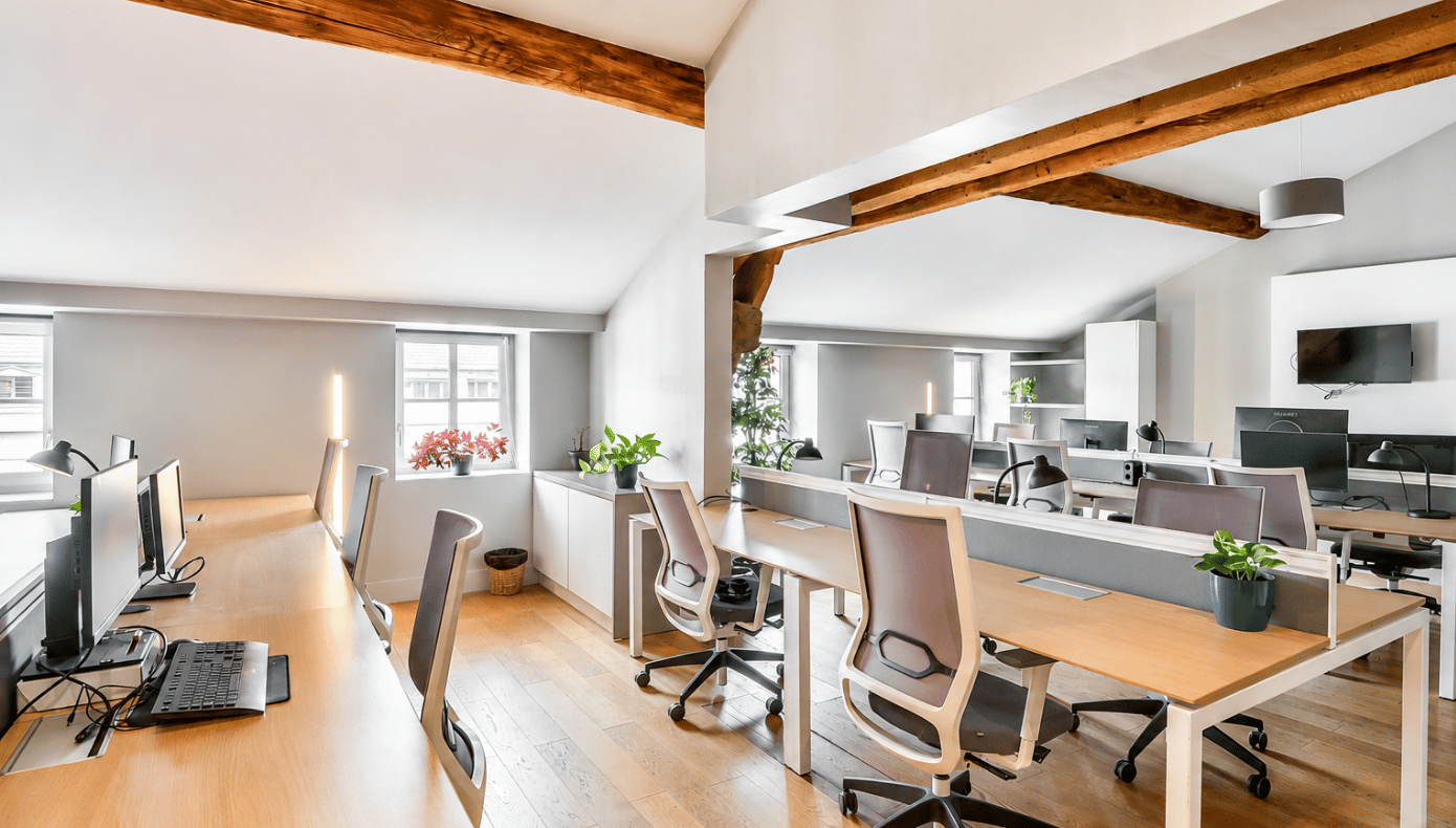 Bureau privatif avec plusieurs postes de travail, écrans, lampes de bureau et poutres en bois apparentes.