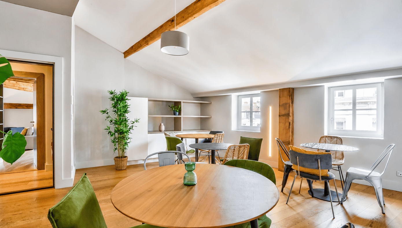 Espace détente aménagé avec tables rondes, assises variées, plantes et fenêtres dans une pièce lumineuse.