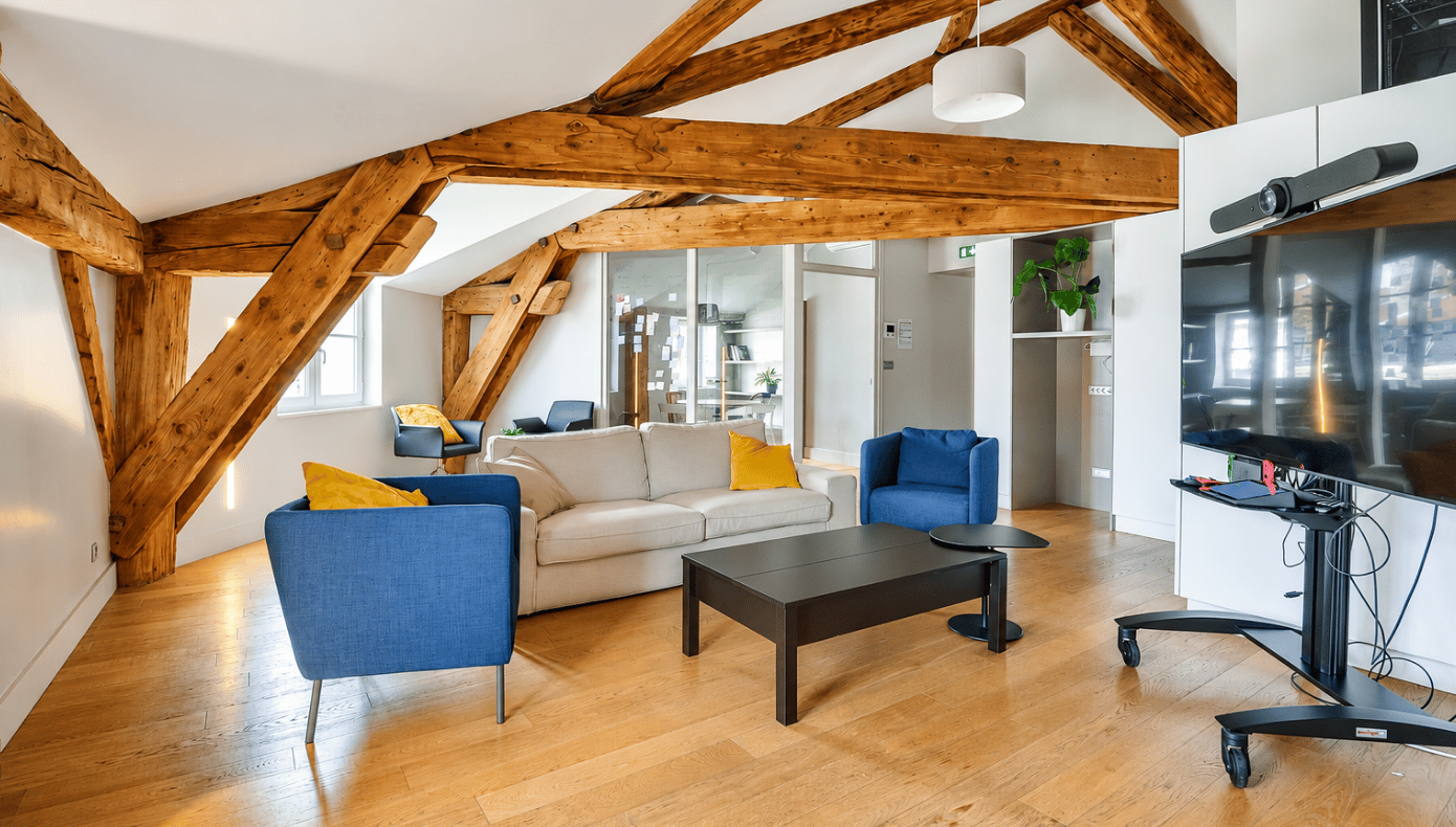 Espace lounge avec canapé, fauteuils, table basse, télévision sur support mobile et poutres en bois apparentes.
