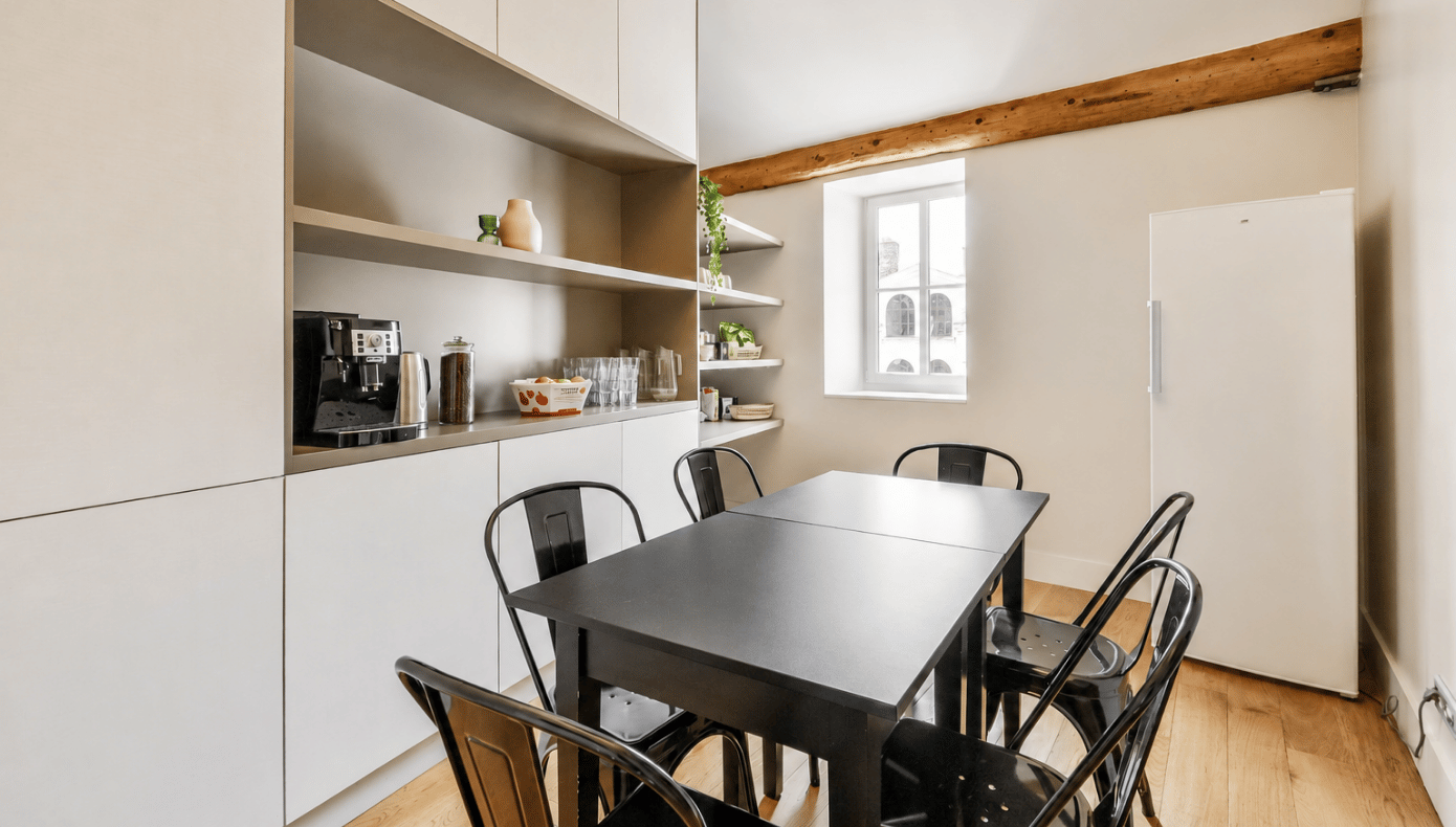 Espace repas avec grande table noire, six chaises, machine à café, rangements et réfrigérateur.
