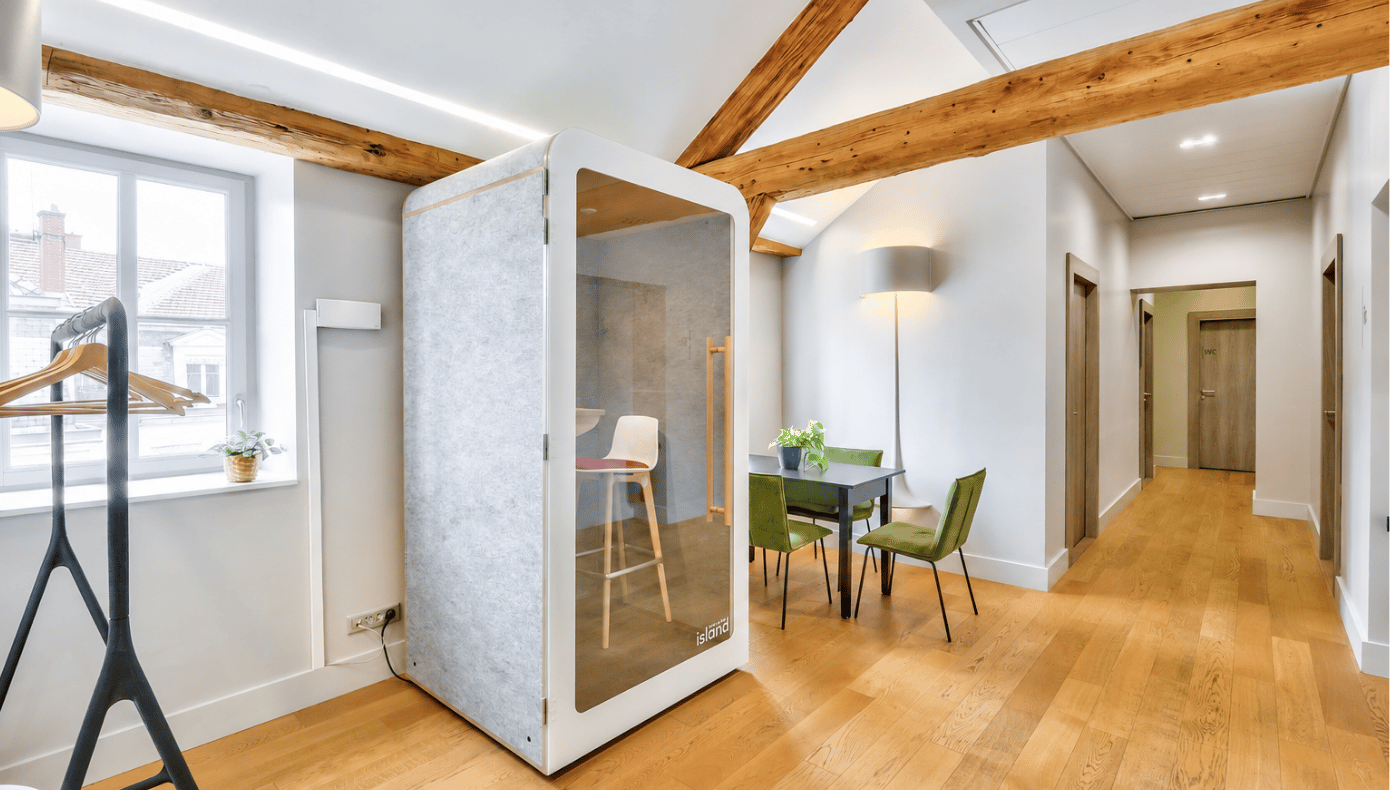 Phonebox vitrée avec siège, installée près d’une petite table et de chaises vertes dans un espace de travail.