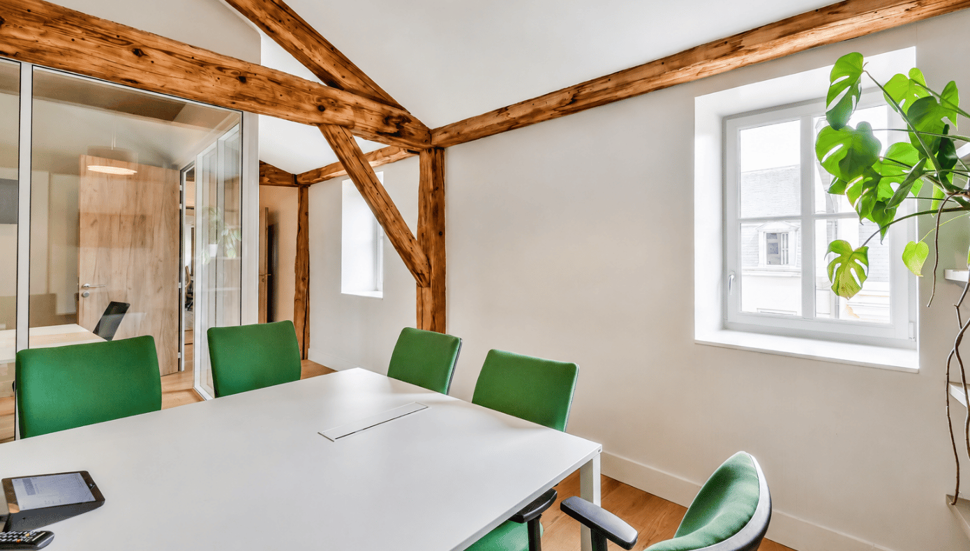 Salle de réunion avec grande table blanche, six chaises vertes, cloisons vitrées et poutres en bois apparentes.