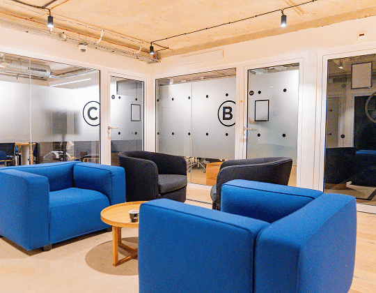 Espace lounge avec fauteuils bleus et noirs, table basse en bois et salles de réunion vitrées en arrière-plan