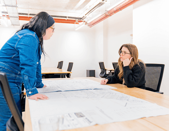 ="Deux femmes en discussion autour de plans d'architecture dans un espace de coworking moderne avec bureaux partagés