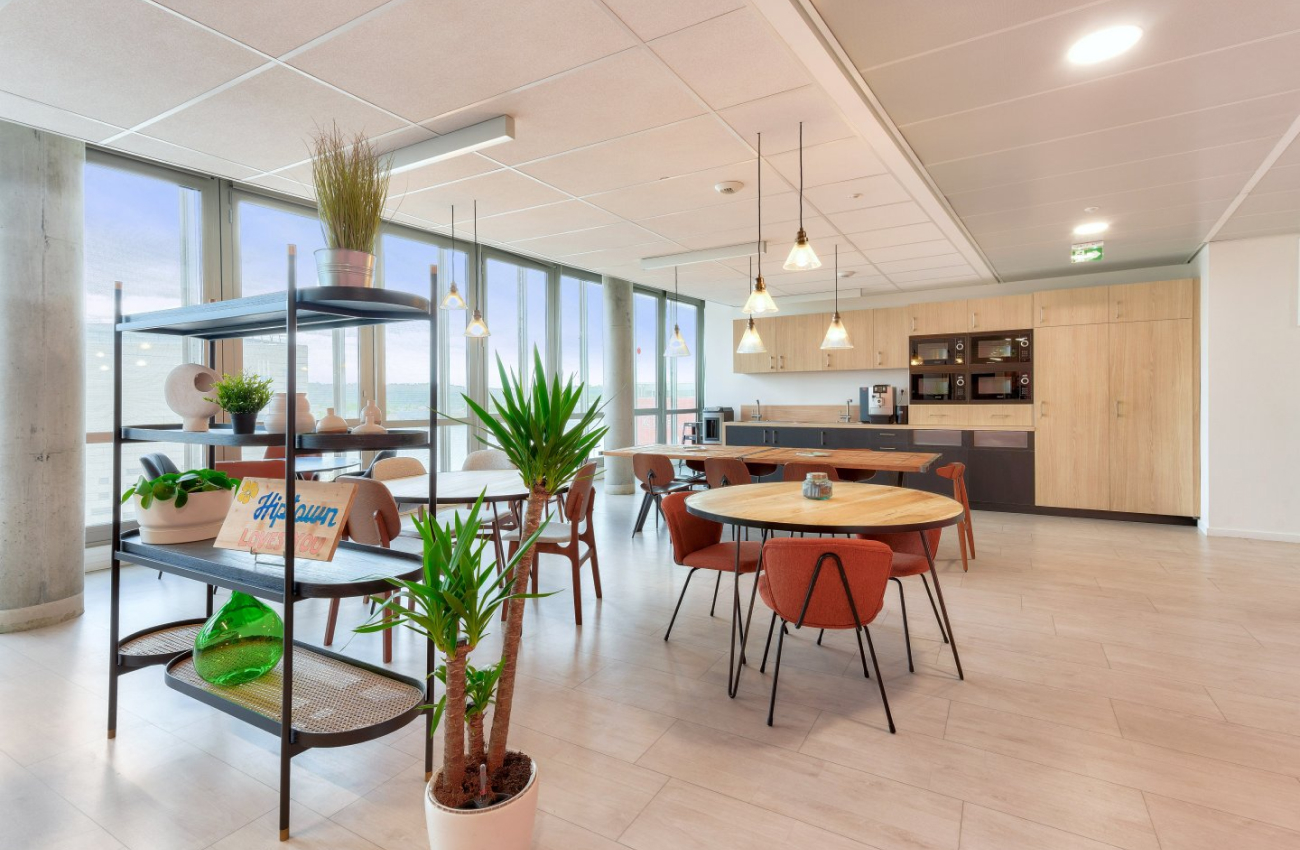 Intérieur lumineux d'un espace de coworking moderne avec des tables en bois, des chaises oranges, des plantes vertes et une cuisine équipée au Quai de Paludate à Bordeaux.