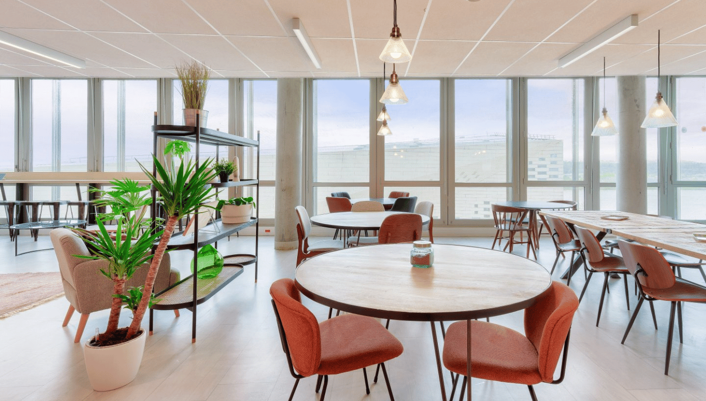 Espace commun avec tables, assises, plantes et grandes baies vitrées dans les bureaux Hiptown à Bordeaux.
