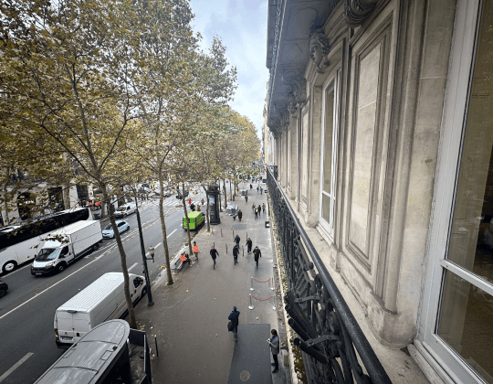 Vue depuis un balcon sur une rue animée de Paris Madeleine, bordée d'arbres et d'immeubles haussmanniens, illustrant l'effervescence du quartier