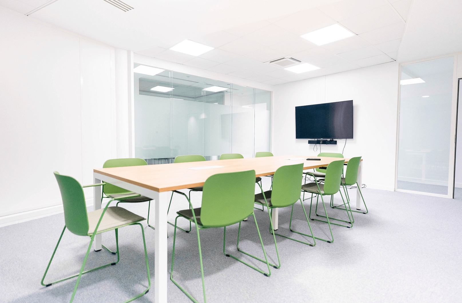 Salle de réunion lumineuse et moderne avec une table en bois et des chaises vertes, équipée d'un écran mural et entourée de parois vitrées