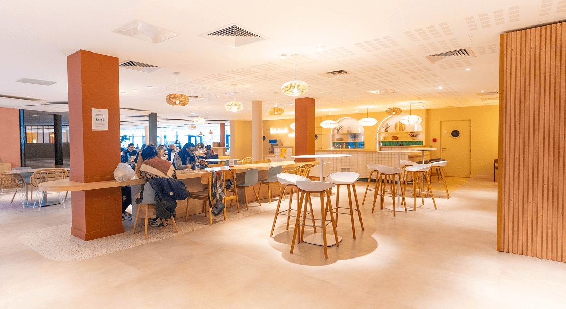 Cafétaria lumineuse et chaleureuse avec un design moderne, bar central, tables hautes et assises confortables, situé dans un bureau flexible Hiptown à Pont Saint-Cloud