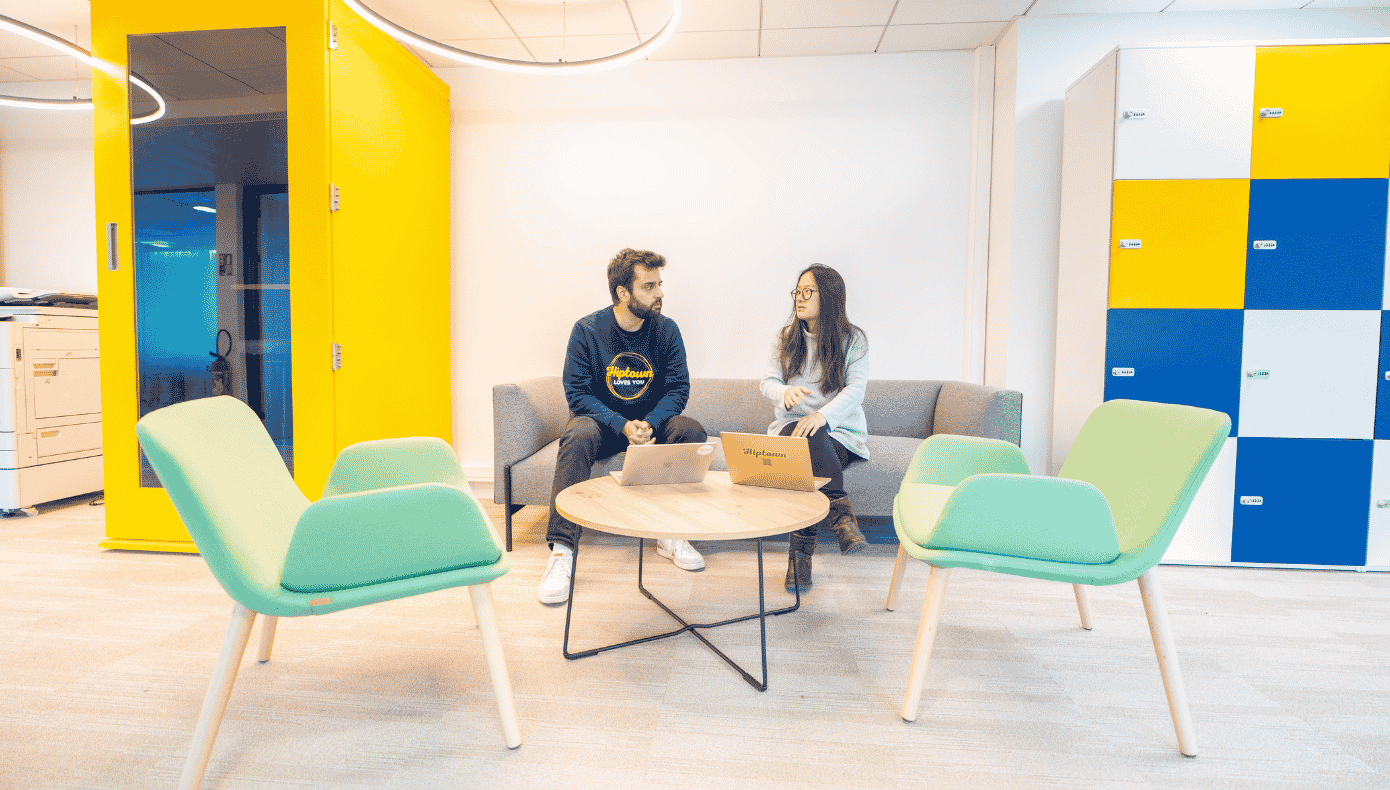 Deux personnes échangent dans un espace détente avec canapé, fauteuils, table basse et casiers colorés.