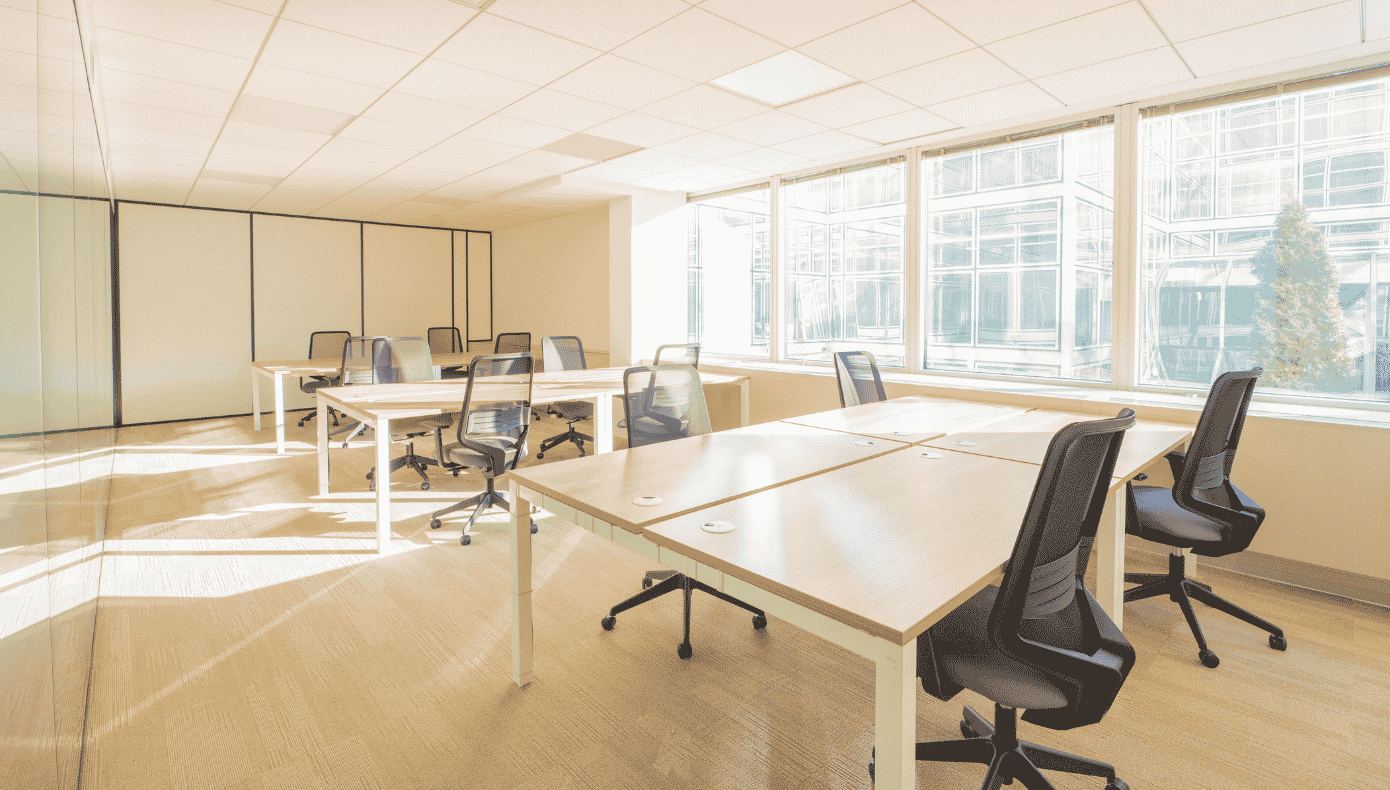 Open space avec plusieurs bureaux, chaises à roulettes et grandes fenêtres donnant sur un bâtiment voisin.