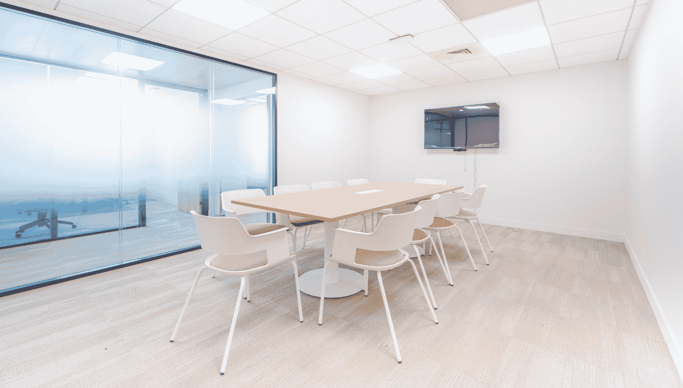 Salle de réunion avec grande table, chaises blanches, écran mural et cloison vitrée.