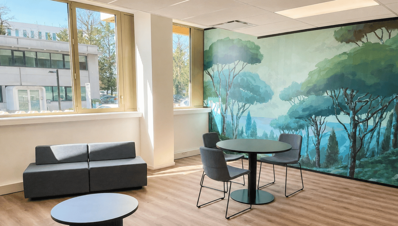 Espace lounge Hiptown à Mérignac avec table ronde, assises grises, grandes fenêtres et fresque végétale.