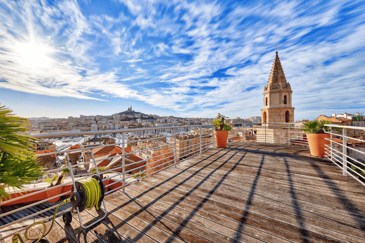 Panoramic view of Marseille's Old Port, with the Basilica of Notre-Dame-de-la-Garde in the background, under a partly cloudy blue sky.