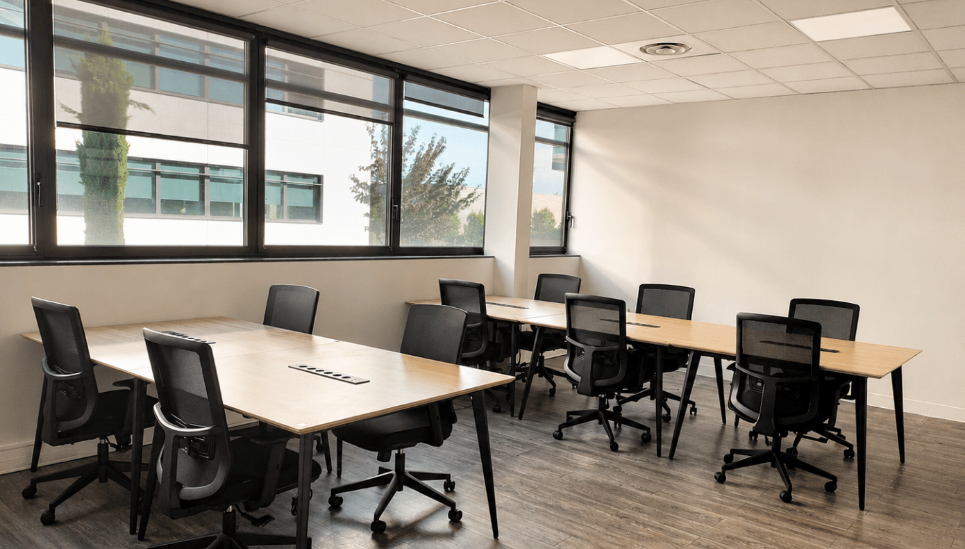 Grand open space organisé en plusieurs îlots de bureaux, avec sièges mobiles et larges fenêtres latérales.