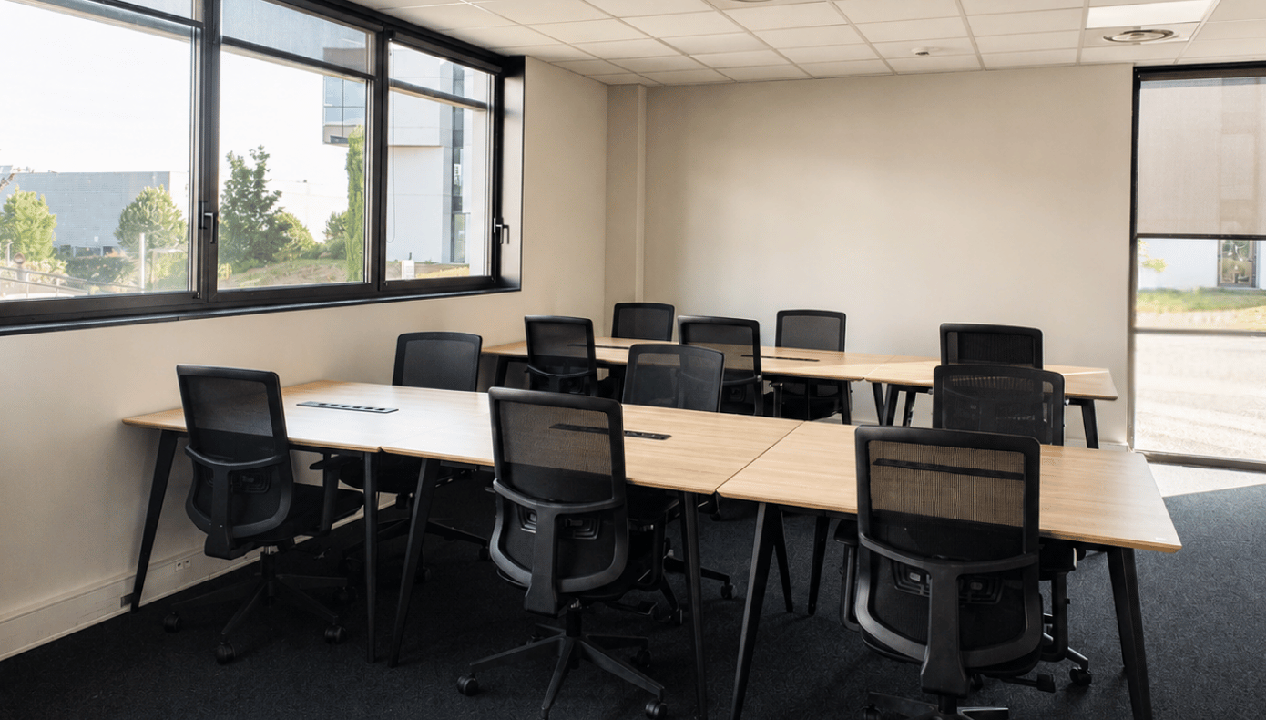 Open space avec plusieurs tables de travail, sièges de bureau et larges fenêtres donnant sur les bâtiments voisins.