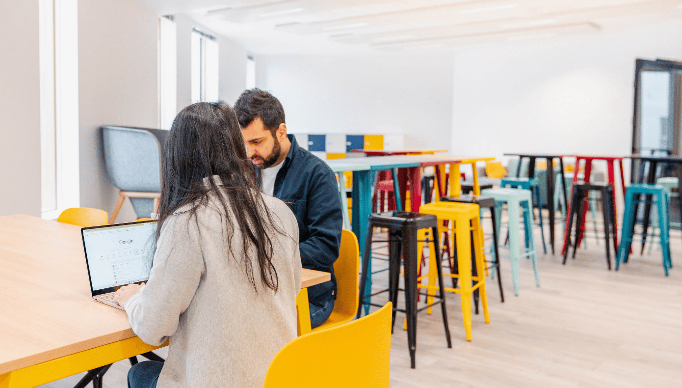 Salle de repas à Pantin avec tables hautes colorées et deux personnes travaillant sur ordinateur portable.